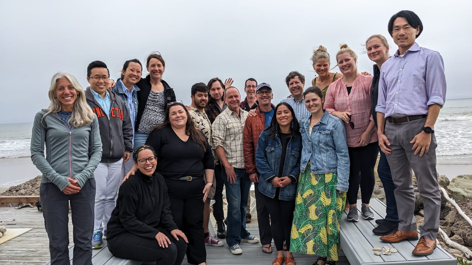 18 people posing for a group picture on a cloudy day in front of a beach