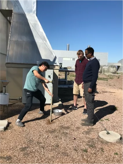 A group of three men stands outside installing a dust catcher with a rubber mallet, stainless steel rod, and a cake pan filled with dark-colored marbles.