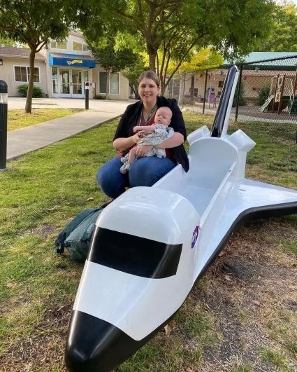 Woman with baby in arms crouches next to model space shuttle