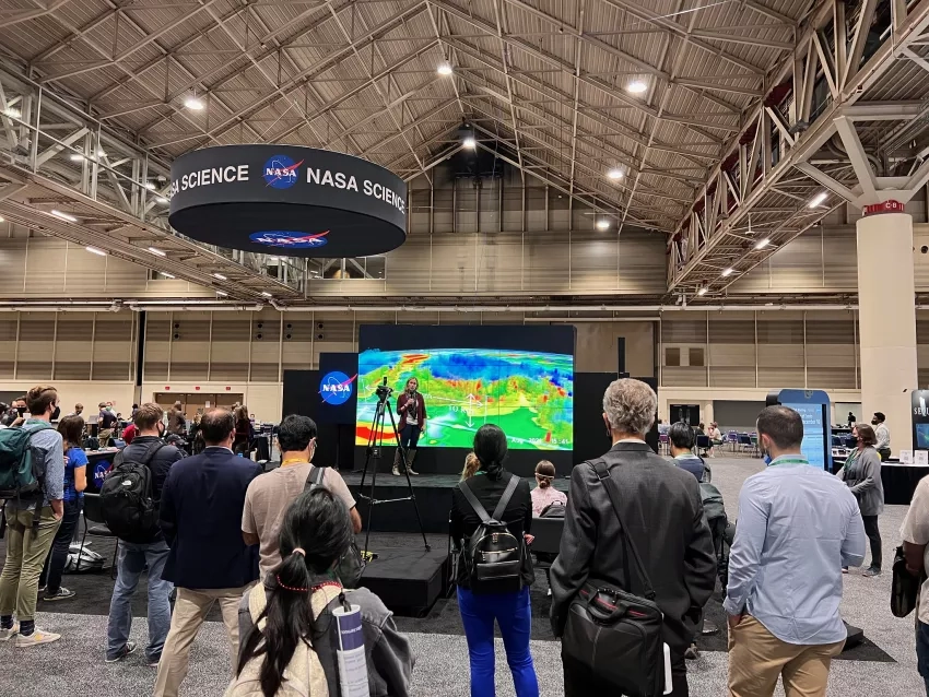 A woman named Dalia Kirschbaum stands on the stage in front of a large screen displaying data visualizations, surrounded by people watching,at the AGU 2021 Fall Meting.