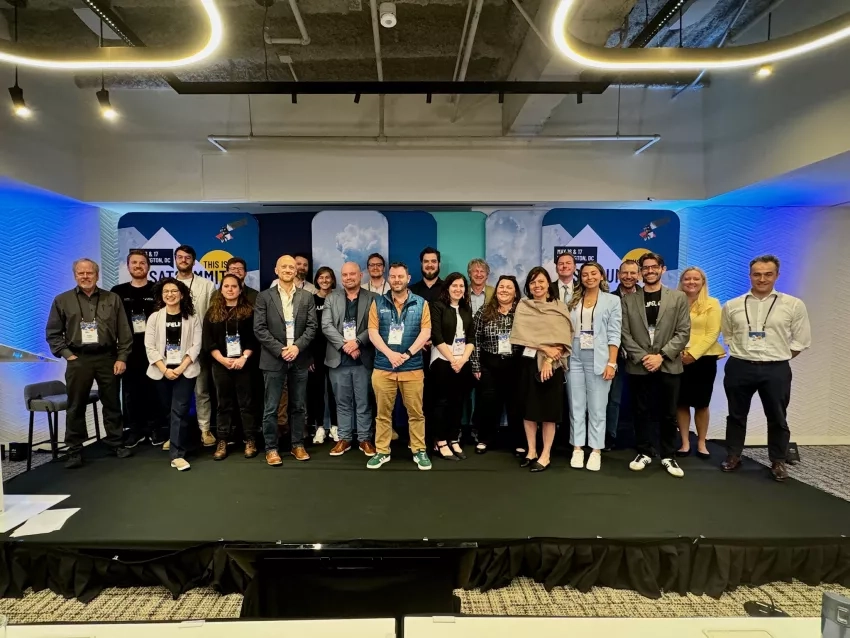 A group of about 20 adults pose for a group photo, smiling. On the wall behind them is a decorative background of clouds, blue light, and imagery of satellites.