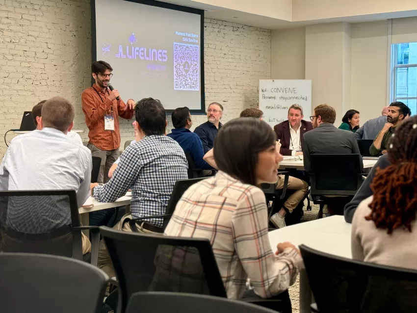 Jeff Pituch stands at the front of a room giving a presentation at the Scientist Speed Dating event. Groups of people sit at tables, eager to participate in this networking event.] 