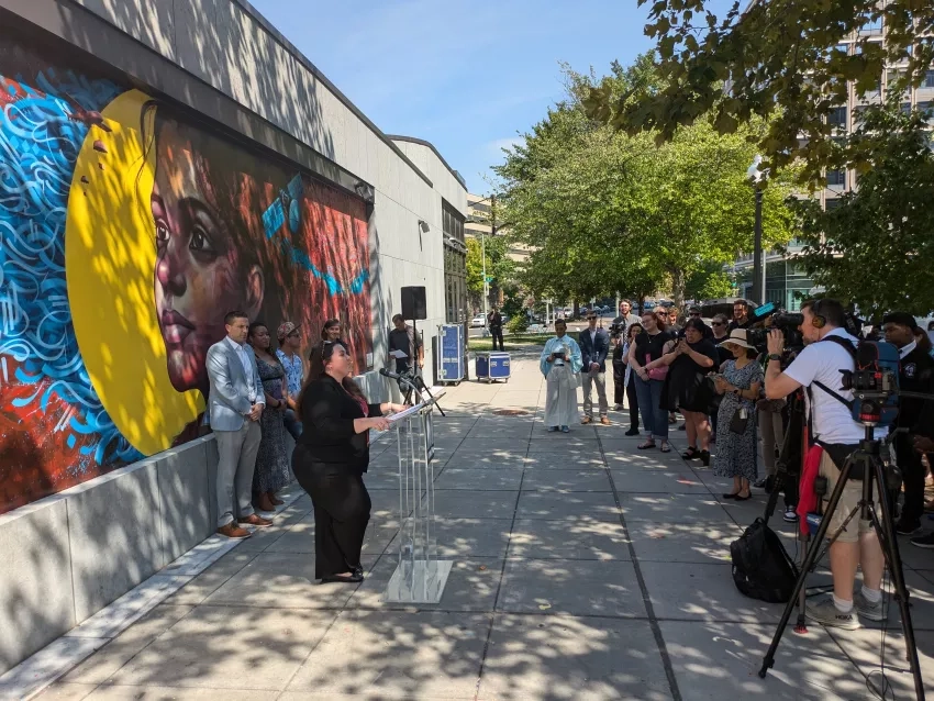 Shanna McClain speaks at a podium in front of the mural on a bright sunny day, with people involved in developing the mural on the left and a large crowd of onlookers and cameramen on the right who are listening to her speech with rapt attention.