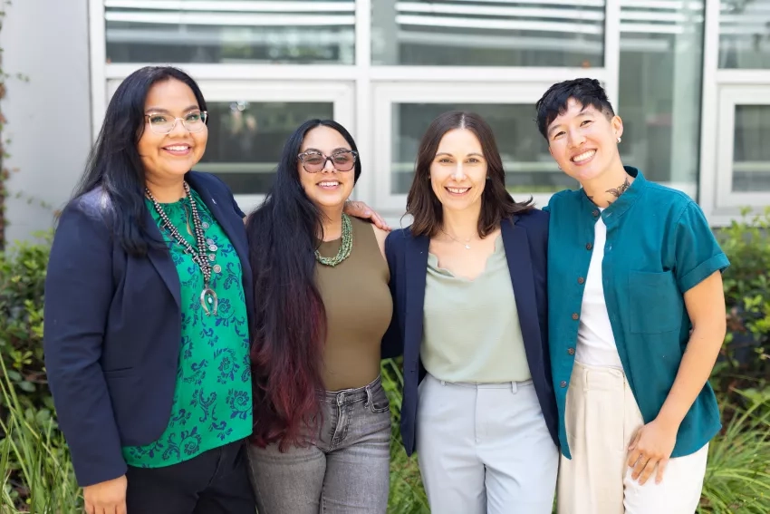 The Indigenous Peoples Initiative team, four individuals, stand in front of greenery and smile at the camera.