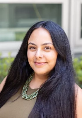 Sativa Cruz, wearing an olive green shirt and a turquoise necklace, smiles at the camera in front of a wall of greenery.