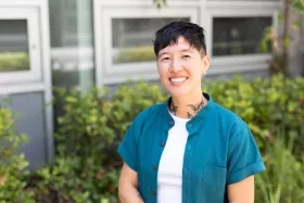 Victoria Ly stands in front of greenery and smiles, wearing a teal button down shirt over a white t-shirt.