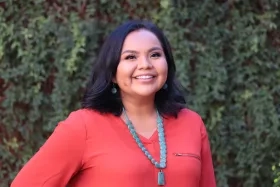 Nikki Tulley, wearing a coral shirt and a turquoise necklace, smiles at the camera in front of a wall of greenery.