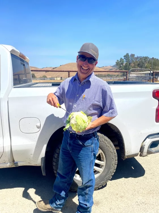 Photo of white man in hat holding a vegetable in a field, smiling.