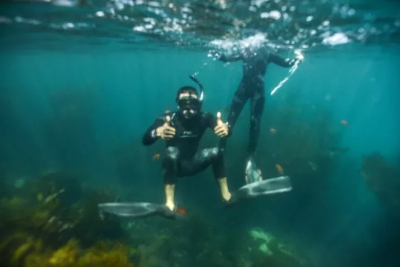 An underwater photo of Ved snorkeling and giving two thumbs up to the camera. Behind him another snorkeler is surfacing.