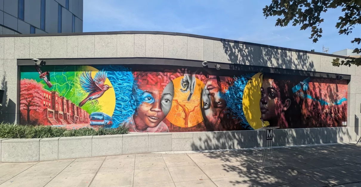 The NASA Lifelines mural is painted with vibrant colors that give feelings of hope and optimism. The mural contains many images that convey the themes of community, humanitarianism, and satellite data. From left to right, the mural includes imagery of a satellite, a DC city street and a metro bus, a bird in flight, two children and a hand pointing at a map of Washington D.C., a woman staring wistfully into the distance, and a group of refugees working together under a satellite map. 