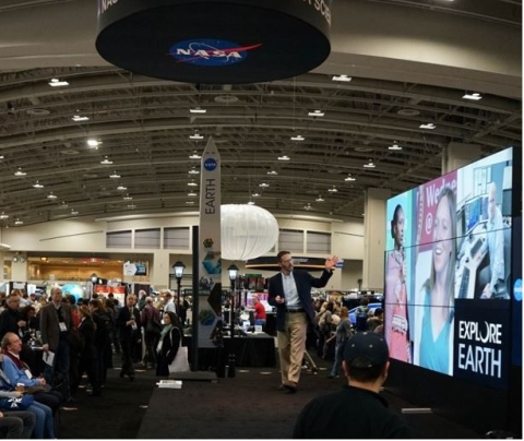 Photo of man on stage gesturing toward a wall of video screens