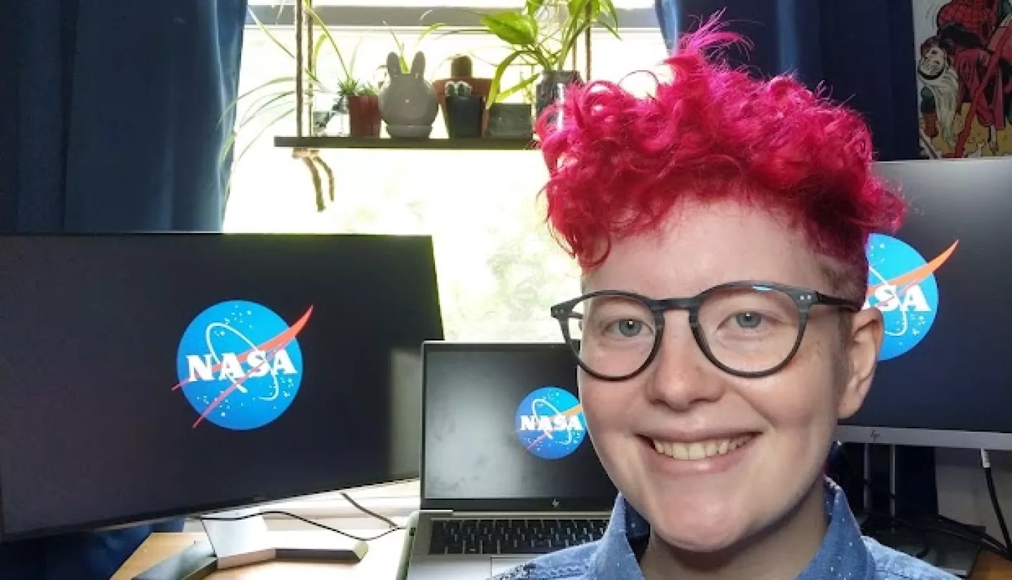 Ila White, who is wearing an astronaut-patterned button-up shirt, glasses, and curly pink hair, smiles in front of desk in their home with NASA meatball displayed on laptop and two monitors on the desk.