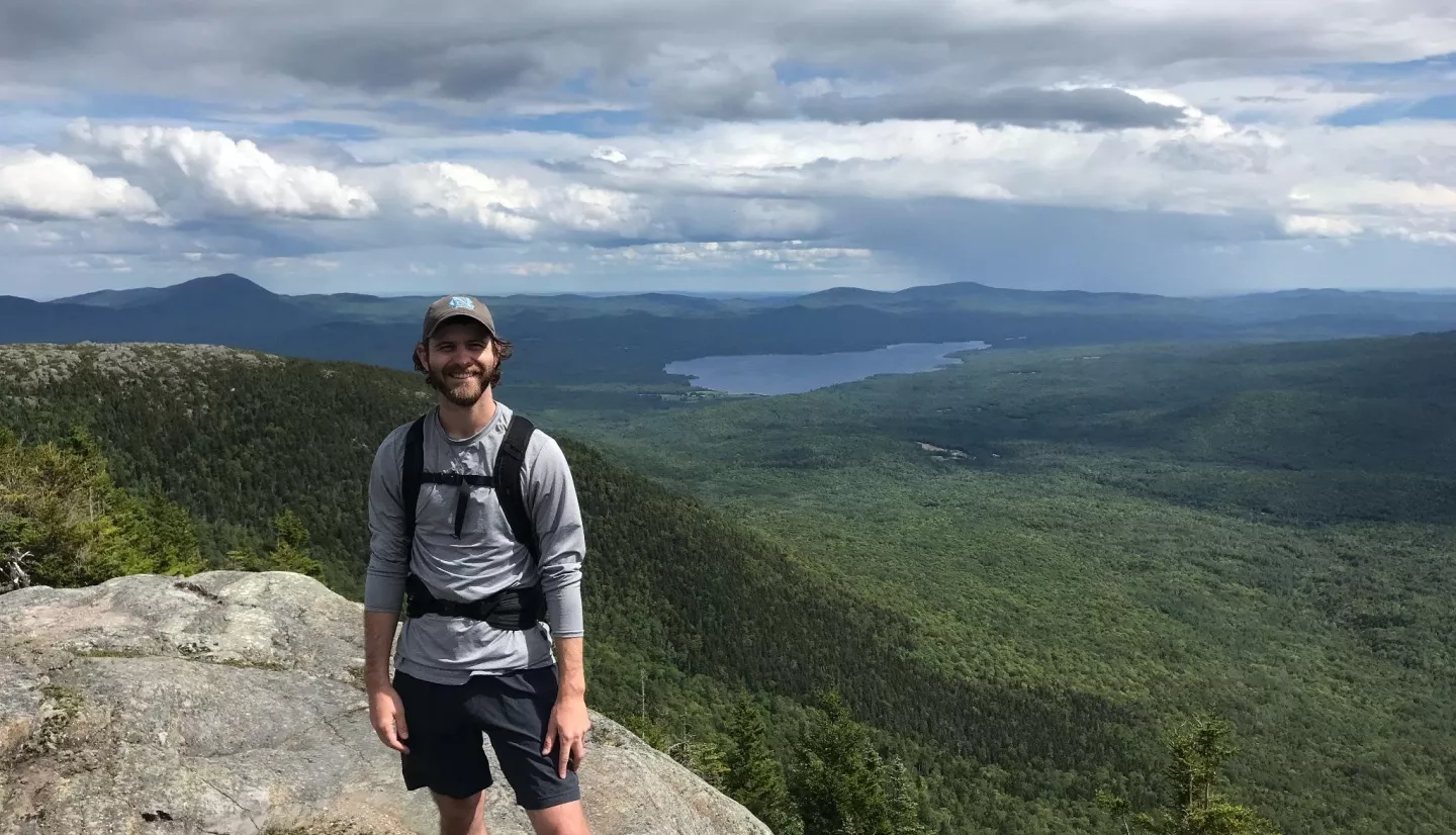 Philip Casey, who wears a hiking backpack and a cap, stands at the top of a rocky mountain with green wooded mountains, a lake in the distance, and clouds hanging overhead.