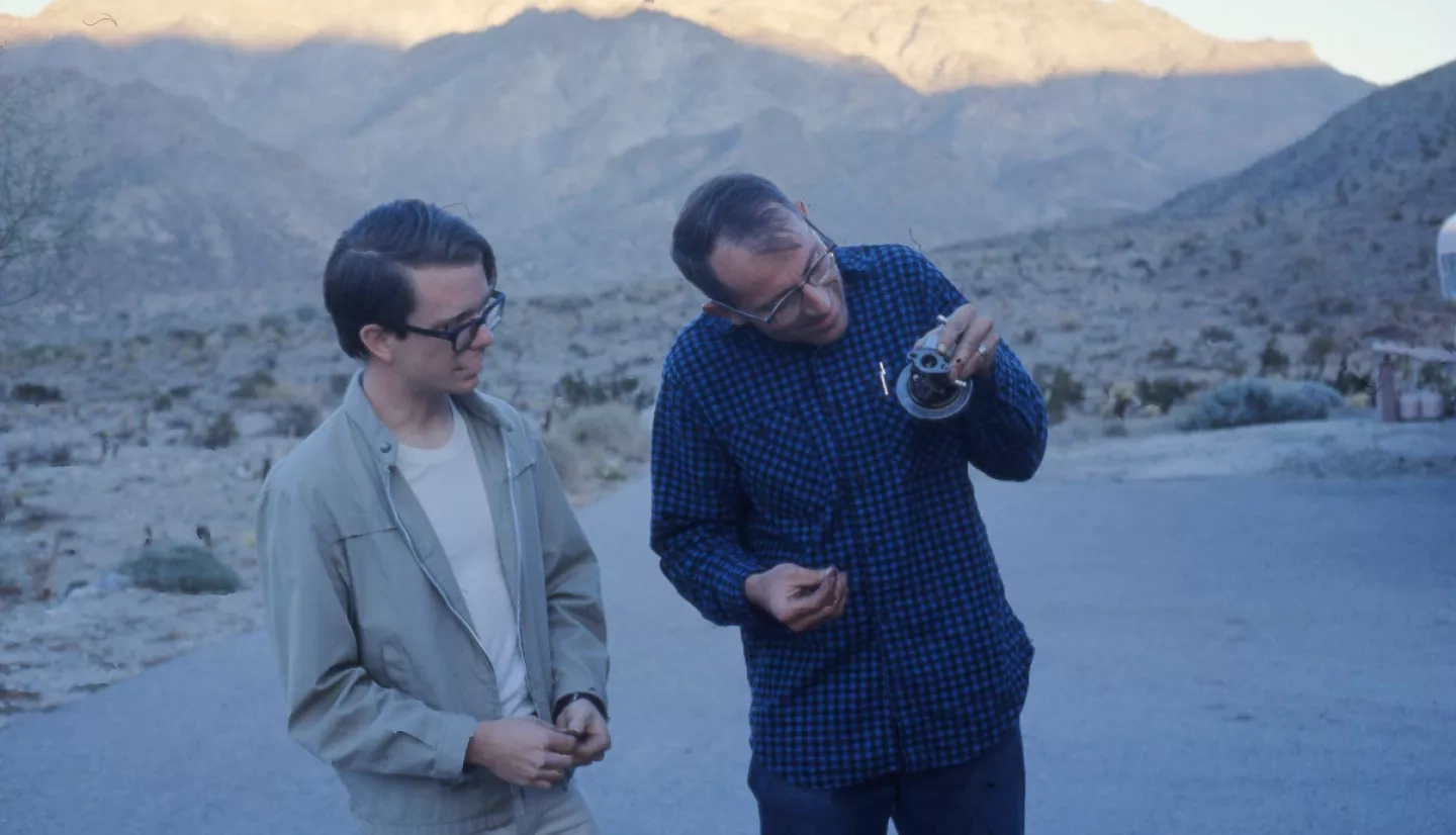 two men inspect a small piece of equipment with mountains in the background