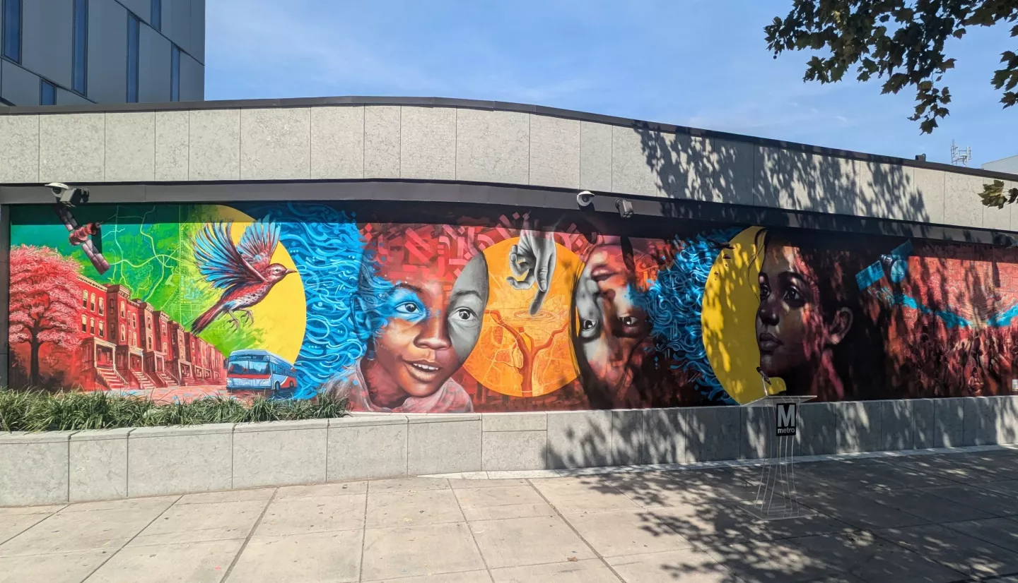 The NASA Lifelines mural is painted with vibrant colors that give feelings of hope and optimism. The mural contains many images that convey the themes of community, humanitarianism, and satellite data. From left to right, the mural includes imagery of a satellite, a DC city street and a metro bus, a bird in flight, two children and a hand pointing at a map of Washington D.C., a woman staring wistfully into the distance, and a group of refugees working together under a satellite map. 