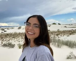 Arlin Arpero stands in a sandy desert with a cloudy sky. Arlin  looks at the camera while facing the left, and wears glasses and a grey NASA Artemis t-shirt.