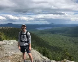 Philip Casey, who wears a hiking backpack and a cap, stands at the top of a rocky mountain with green wooded mountains, a lake in the distance, and clouds hanging overhead.