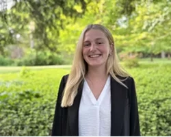 Kameron , wearing a black blazer, smiles at the camera with a grassy field behind her.