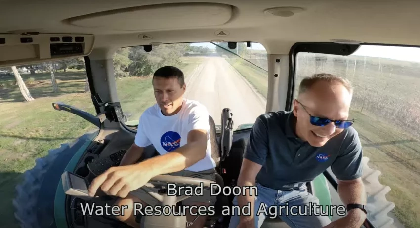 two men in the cab of a tractor with one pointing at fields as they drive along